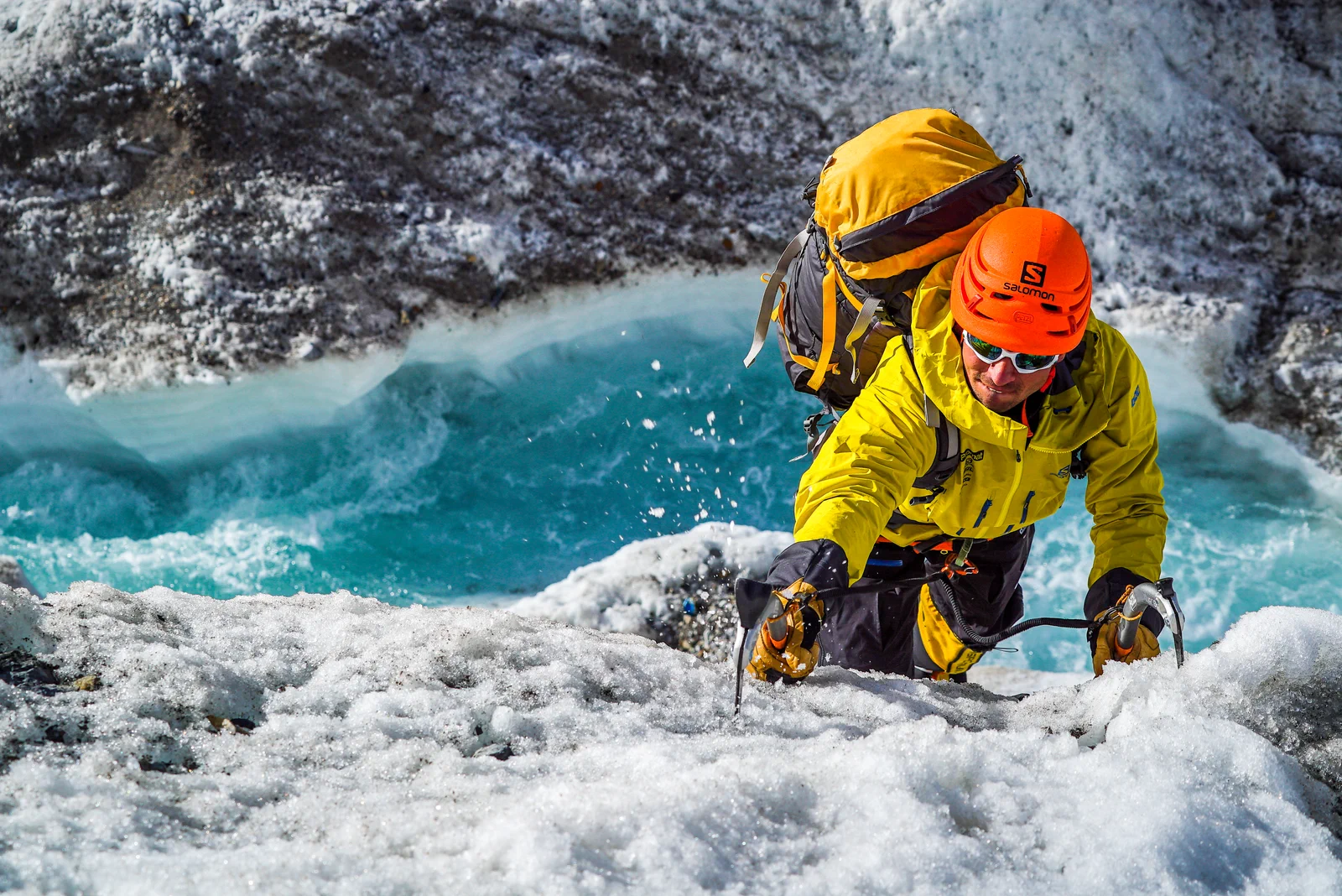 A climber in a yellow jacket and orange helmet crossing a glacier beside a turquoise meltwater channel