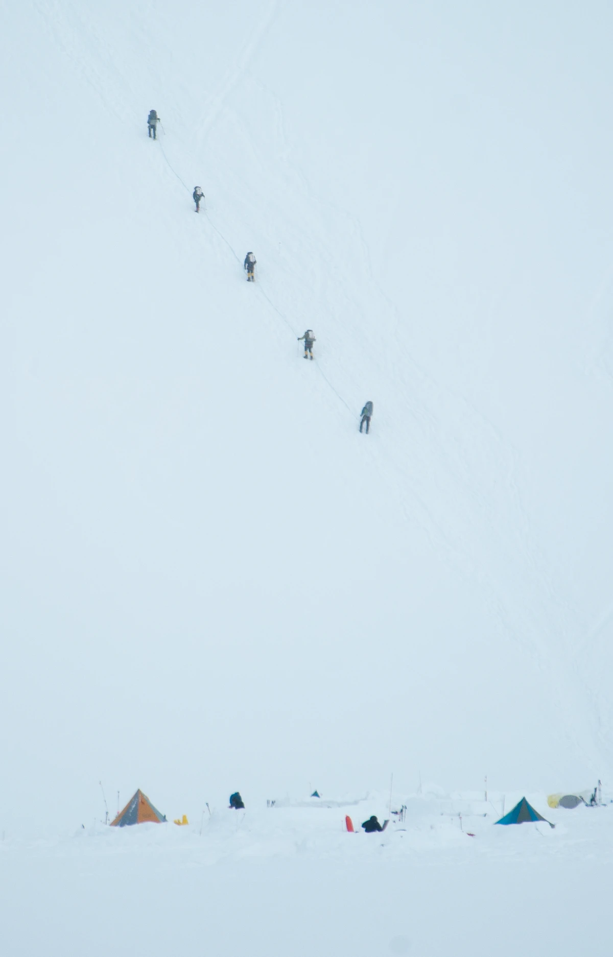 A line of climbers ascending a steep, pale snow slope above a small camp of tents