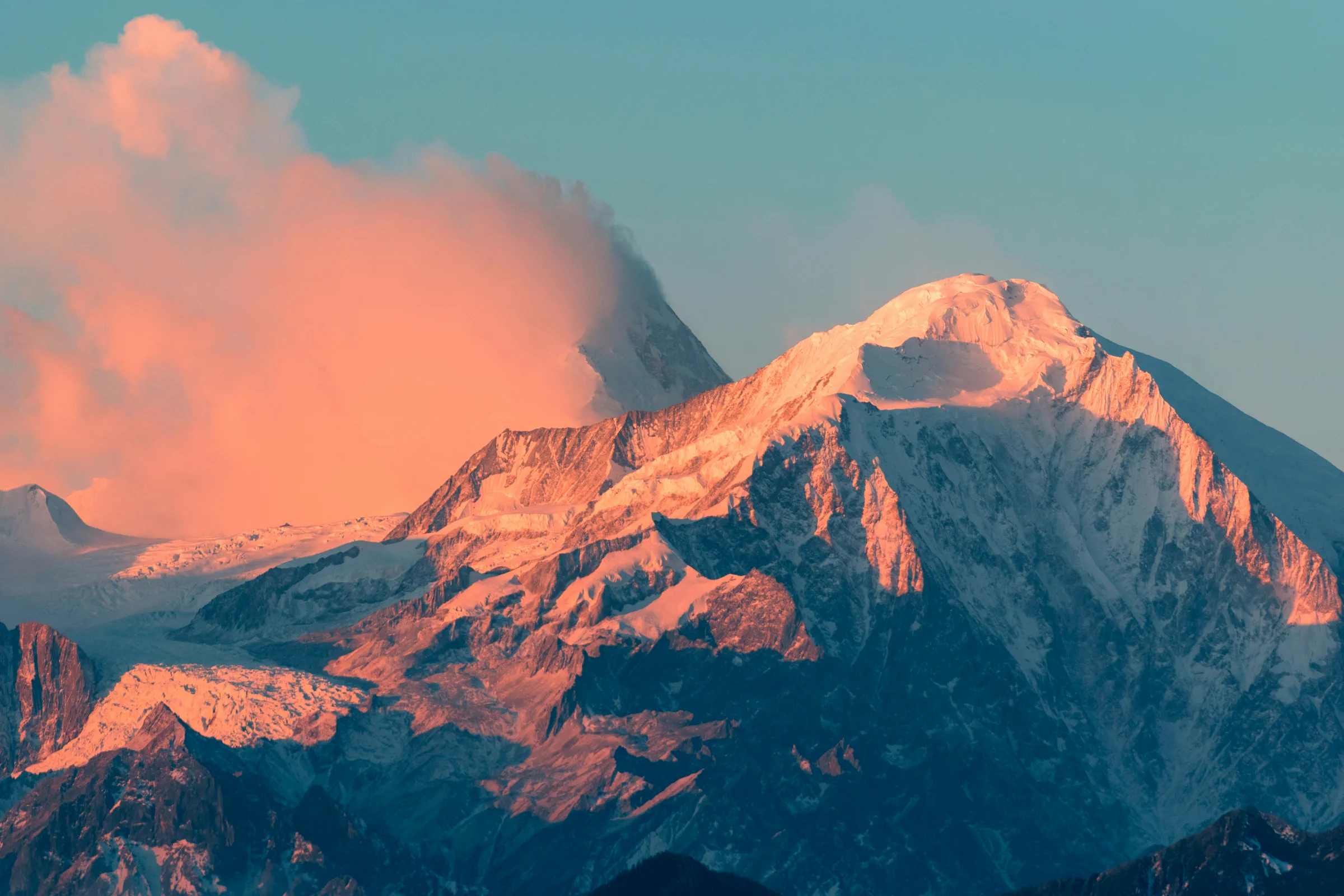 A snow summit glowing orange as first light reaches it, with a plume of cloud drifting beside it